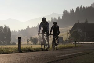 Zwei Radfahrer fahren auf einer Straße vor Bergkulisse