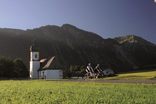 Zwei Radfahrer vor Kirche und Berglandschaft
