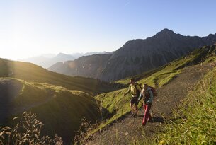 Wanderer auf Bergpfad mit Blick auf grüne Hügel und Gipfel im Sonnenlicht