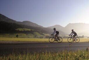 Zwei Radfahrer auf Asphaltstraße vor grüner Landschaft mit Bergen im Hintergrund