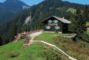 Hütte auf grüner Bergwiese vor bewaldeten Bergen unter blauem Himmel