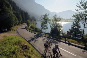 Zwei Radfahrer auf kurviger Straße am See vor Bergkulisse