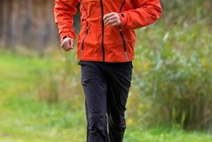 Mann in orangefarbener Jacke und schwarzer Hose joggt auf einem grünen Pfad