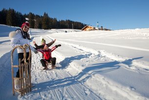 Rodeln im Schnee mit lachenden Personen und Holzhütte im Hintergrund