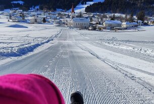 Blick auf verschneite Rodelbahn mit Dorf und Bergen im Hintergrund