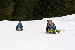 Familie und Kind rodeln im Schnee vor dunklem Wald