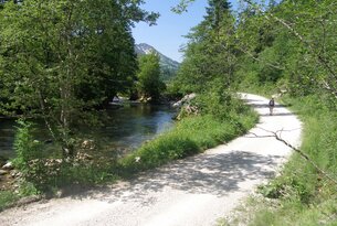 Fluss neben Schotterweg mit Wanderer, Bäumen und Berg im Hintergrund