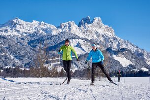 Zwei Langläufer vor verschneiter Bergkulisse an einem sonnigen Tag