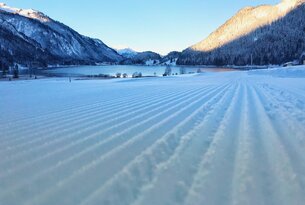 Verschneite Ebene mit Loipen vor See und Bergen unter blauem Himmel
