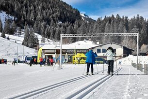 Langläufer auf Loipe vor Gebäude mit Schriftzug SV Nesselwängle, bewaldete Berge im Hintergrund