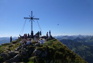 Gipfelkreuz auf Bergspitze mit Menschen unter blauem Himmel