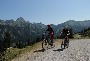 Two mountain bikers ride on a gravel road in front of a mountain panorama