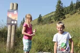 Zwei Kinder stehen an einem Informationsschild vor einer Berglandschaft