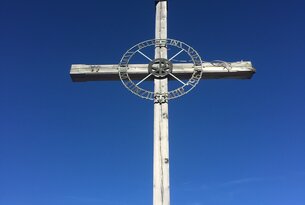 Hölzernes Gipfelkreuz mit Metallring vor blauem Himmel