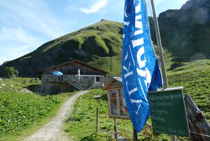 Berghütte mit Weg, Berg im Hintergrund und blau-weiße Fahne im Vordergrund