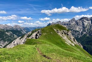Grüner Bergrücken unter blauem Himmel mit Wolken, im Hintergrund Bergkette