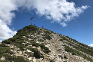 Bergpfad führt zu Gipfelkreuz unter blauem Himmel mit Wolken