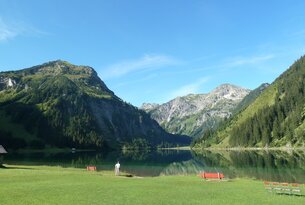 Bergsee mit grüner Wiese, Bänken und Person vor Bergkulisse unter blauem Himmel