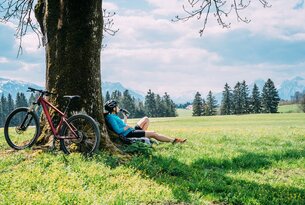 Radfahrerin ruht am Baum mit Blick auf Wiese und Berge