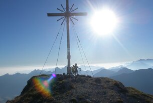 Gipfelkreuz auf Bergspitze mit zwei Personen im Gegenlicht vor Bergpanorama