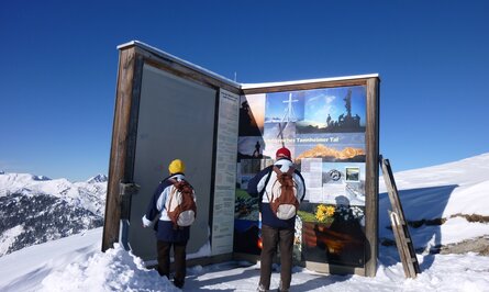 Zwei Wanderer vor einer Informationstafel im Schnee unter blauem Himmel