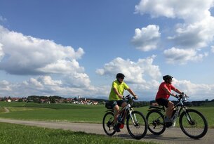 Zwei Personen fahren E-Bike auf asphaltierter Straße, grüne Wiese und blauer Himmel mit Wolken im Hintergrund