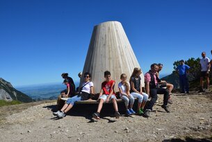 Gruppe sitzt auf Bank vor Holzkonstruktion auf Berggipfel