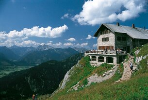 Steinhaus auf Bergspitze mit Menschen, im Hintergrund Bergpanorama unter blauem Himmel mit Wolken
