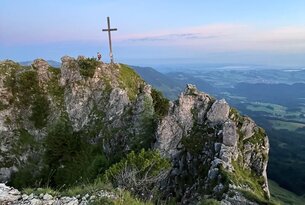 Gipfelkreuz auf felsigem Berggrat mit Blick auf grüne Landschaft im Abendlicht