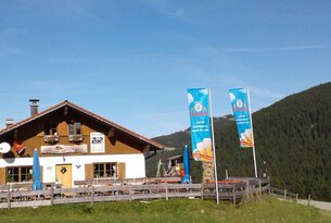 Berghütte mit Biergarten und Fahnen vor bewaldetem Hang unter blauem Himmel