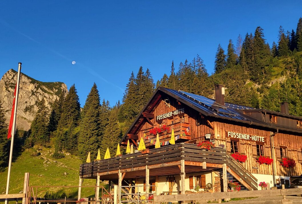 Hölzerne Berghütte mit Balkon vor bewaldetem Hang unter blauem Himmel
