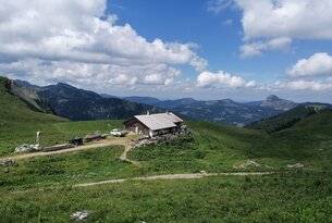 Hütte auf grüner Bergwiese vor Bergpanorama unter blauem Himmel mit Wolken