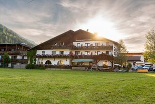 Gasthof mit dunklem Dach, weißen Wänden und Balkonen vor grüner Wiese im Sonnenlicht
