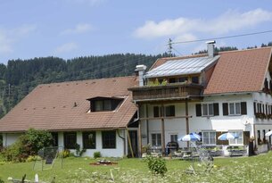 Bauernhaus mit rotem Ziegeldach und Balkon vor bewaldetem Hügel
