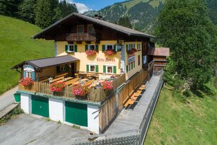 Gelbes Haus mit braunem Dach, grünen Fensterläden und Blumenkästen vor Bergkulisse