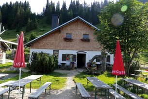 Hütte mit Holzdach und weißen Wänden vor bewaldetem Hang mit Tischen und Bänken davor