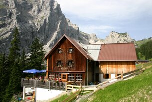 Hütte vor Bergmassiv mit Terrasse und blauem Sonnenschirm