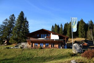 Holzhaus mit bayerischer Flagge auf grüner Wiese unter blauem Himmel