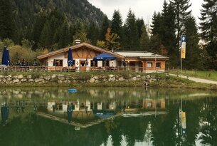 Holzhütte am See mit Spiegelung im Wasser vor bewaldetem Hintergrund