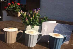 Drei Cappuccinos auf Holztisch, daneben Topf mit Enzian und Speisekarte der Zipfelsalpe