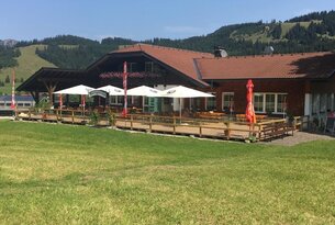 Gasthof mit Terrasse und Sonnenschirmen vor grüner Wiese und bewaldeten Hügeln