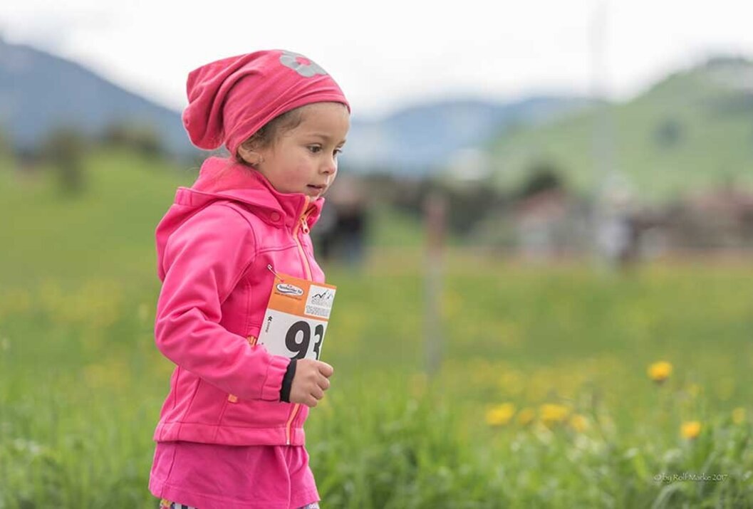 Mädchen mit rosa Jacke und Mütze hält Startnummer in der Hand, Wiese im Hintergrund