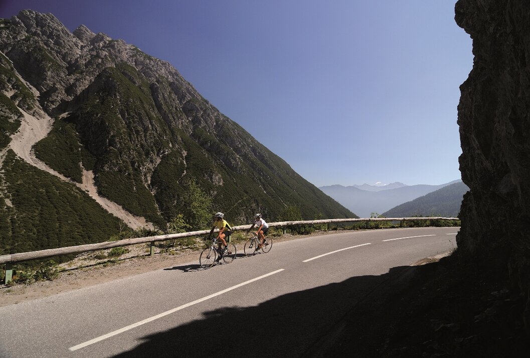 Zwei Radfahrer auf Bergstraße vor Bergkulisse unter blauem Himmel