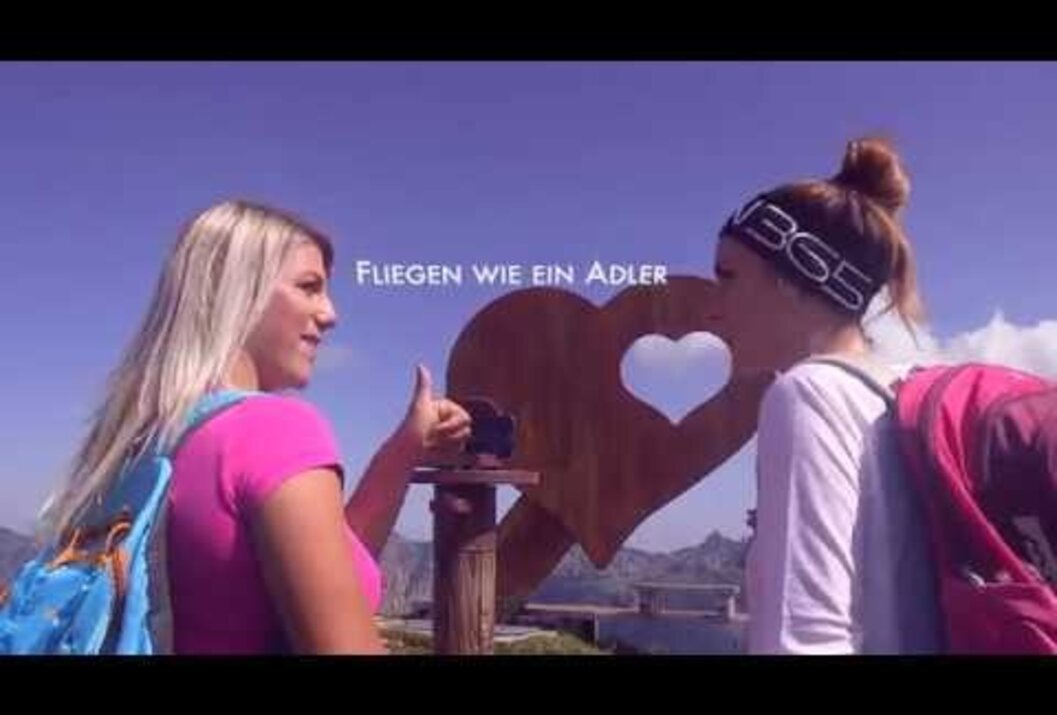 Zwei Frauen vor Herzskulptur mit Bergblick und Text "Fliegen wie ein Adler"