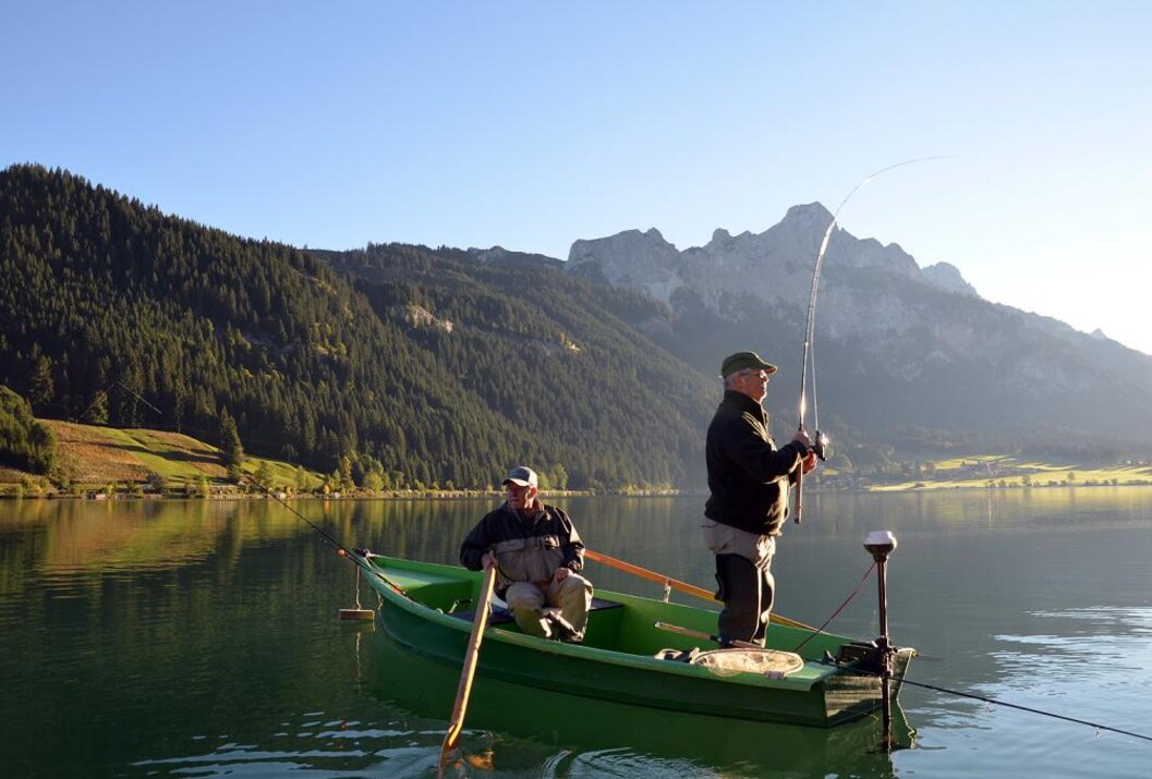 Zwei Männer angeln in einem grünen Boot auf einem See vor Bergkulisse