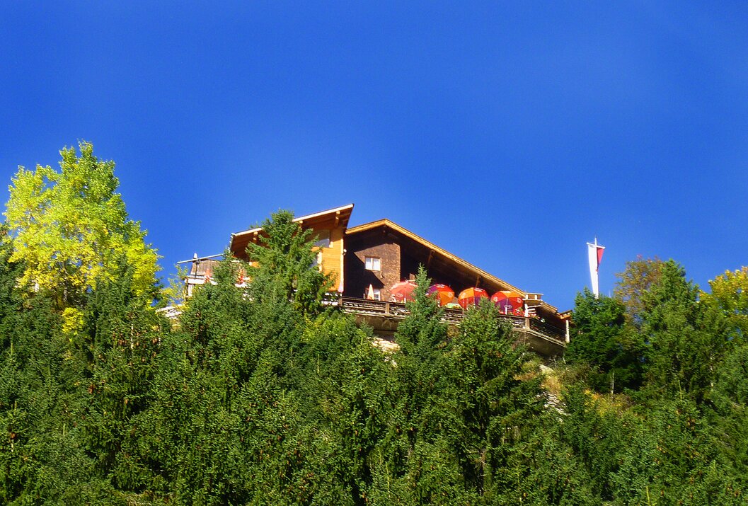 Holzhaus auf einem Hügel, umgeben von Bäumen unter blauem Himmel