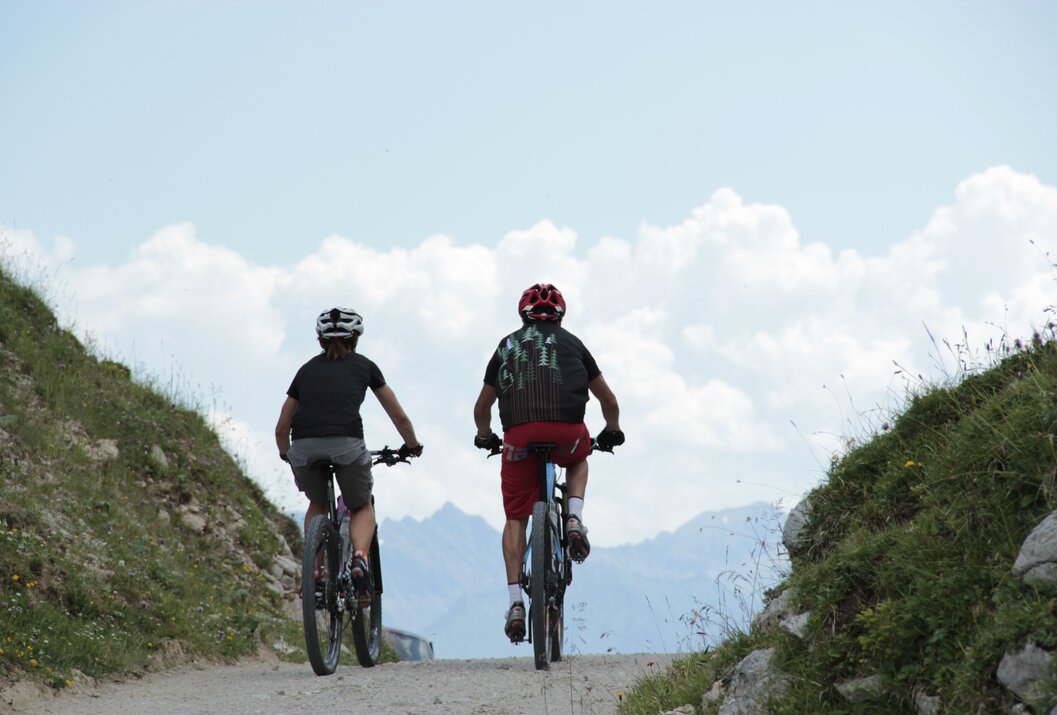 Zwei Mountainbiker fahren bergauf auf einem Schotterweg vor Bergkulisse