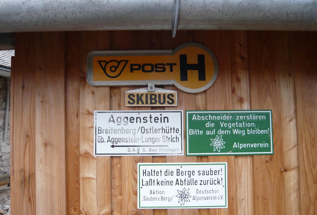Schilder mit Hinweisen zu Post, Skibus, Aggenstein, Naturschutz und Sauberkeit an Holzwand