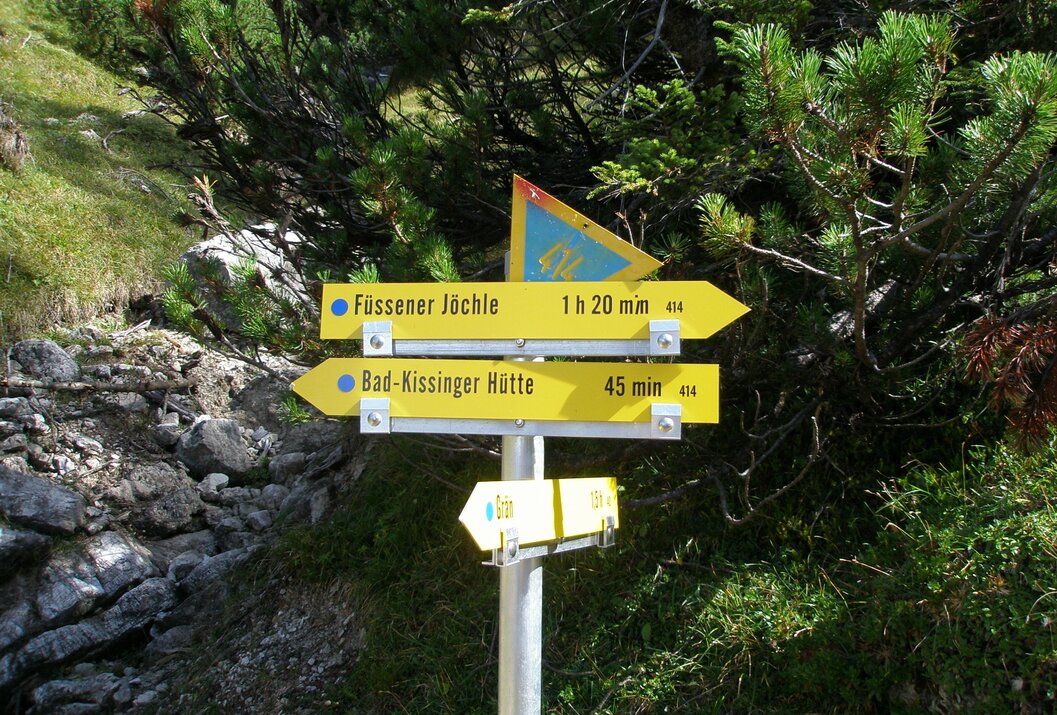 Gelbe Wegweiser mit Zielen und Zeitangaben vor Bäumen und Felsen