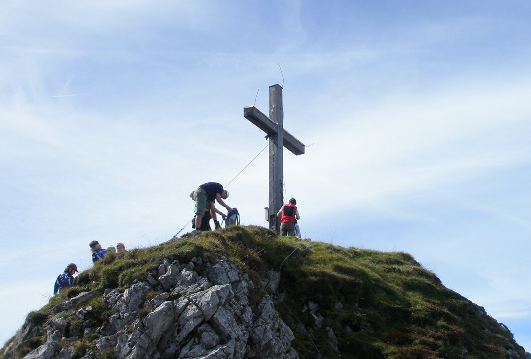 Gipfelkreuz auf Bergspitze mit Menschen und Felsen unter blauem Himmel